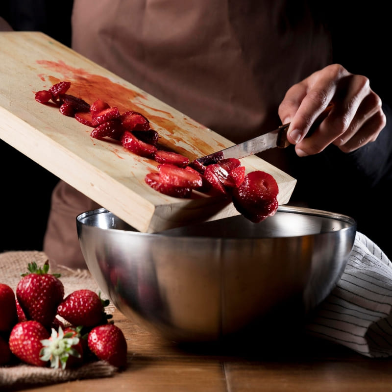 Person slicing fresh strawberries onto a wooden cutting board and transferring them into a stainless steel mixing bowl, with whole strawberries nearby.
