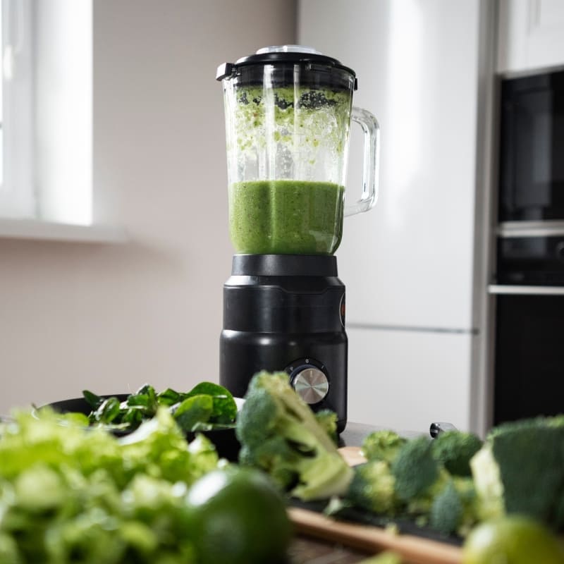 Green smoothie blending in a countertop blender surrounded by fresh vegetables including spinach, broccoli, lettuce, and lime in a bright modern kitchen ā perfect for a healthy detox or green smoothie recipe.