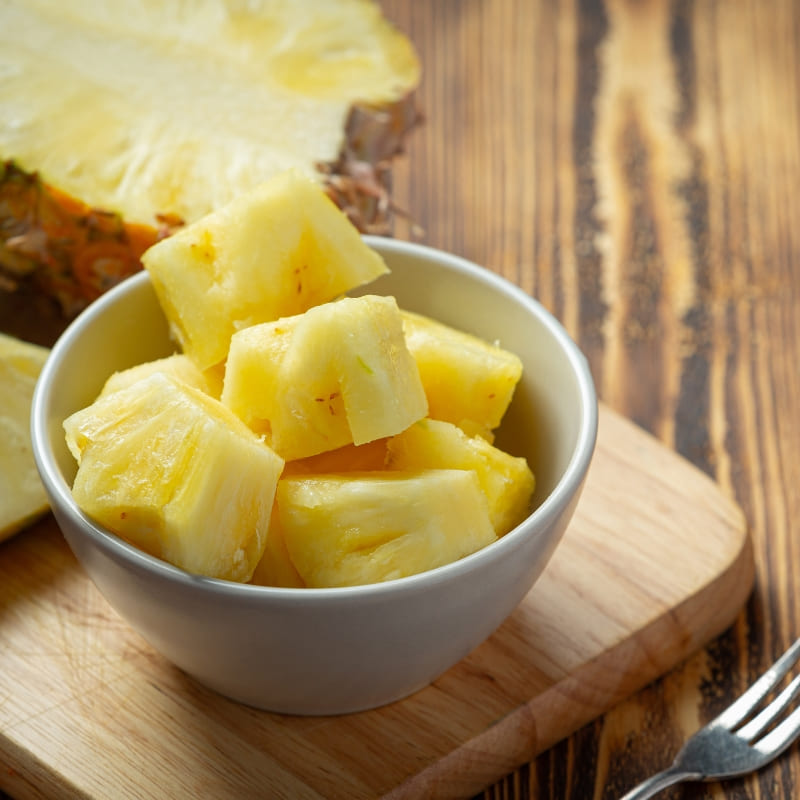 Fresh pineapple chunks in a white bowl on a wooden cutting board with a halved pineapple in the background ā tropical fruit ready for smoothies or healthy snacking.