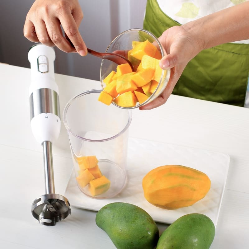 Person preparing a mango smoothie by pouring fresh mango chunks into a blender cup, with a peeled mango, whole mangoes, and an immersion blender on a white kitchen counter ā healthy smoothie preparation with tropical fruit.